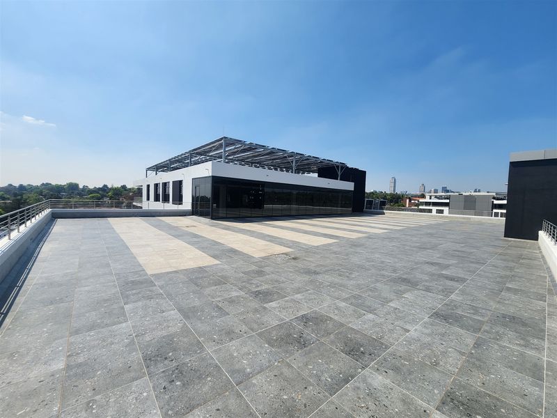 Expansive rooftop terrace featuring modern tiled flooring, metal safety railings, and a large glass-fronted office structure with integrated solar panel roofing.