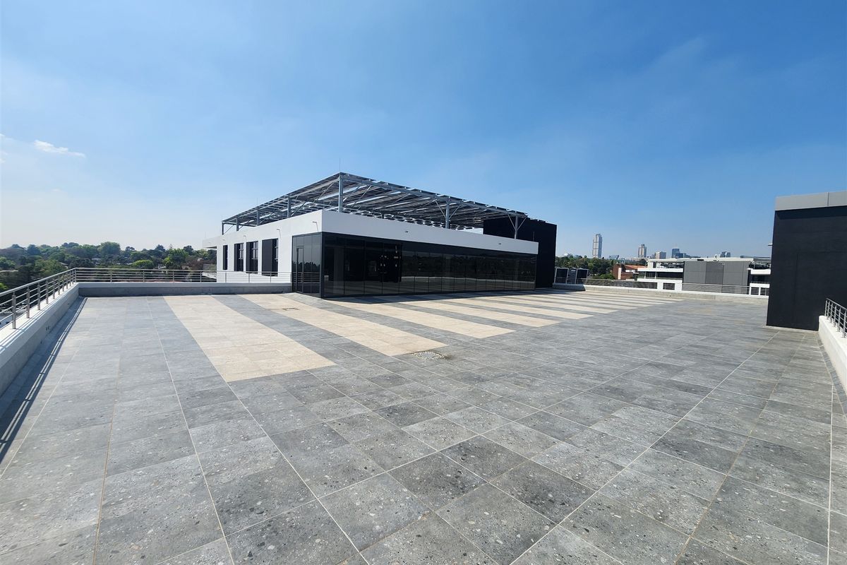 Expansive rooftop terrace featuring modern tiled flooring, metal safety railings, and a large glass-fronted office structure with integrated solar panel roofing.