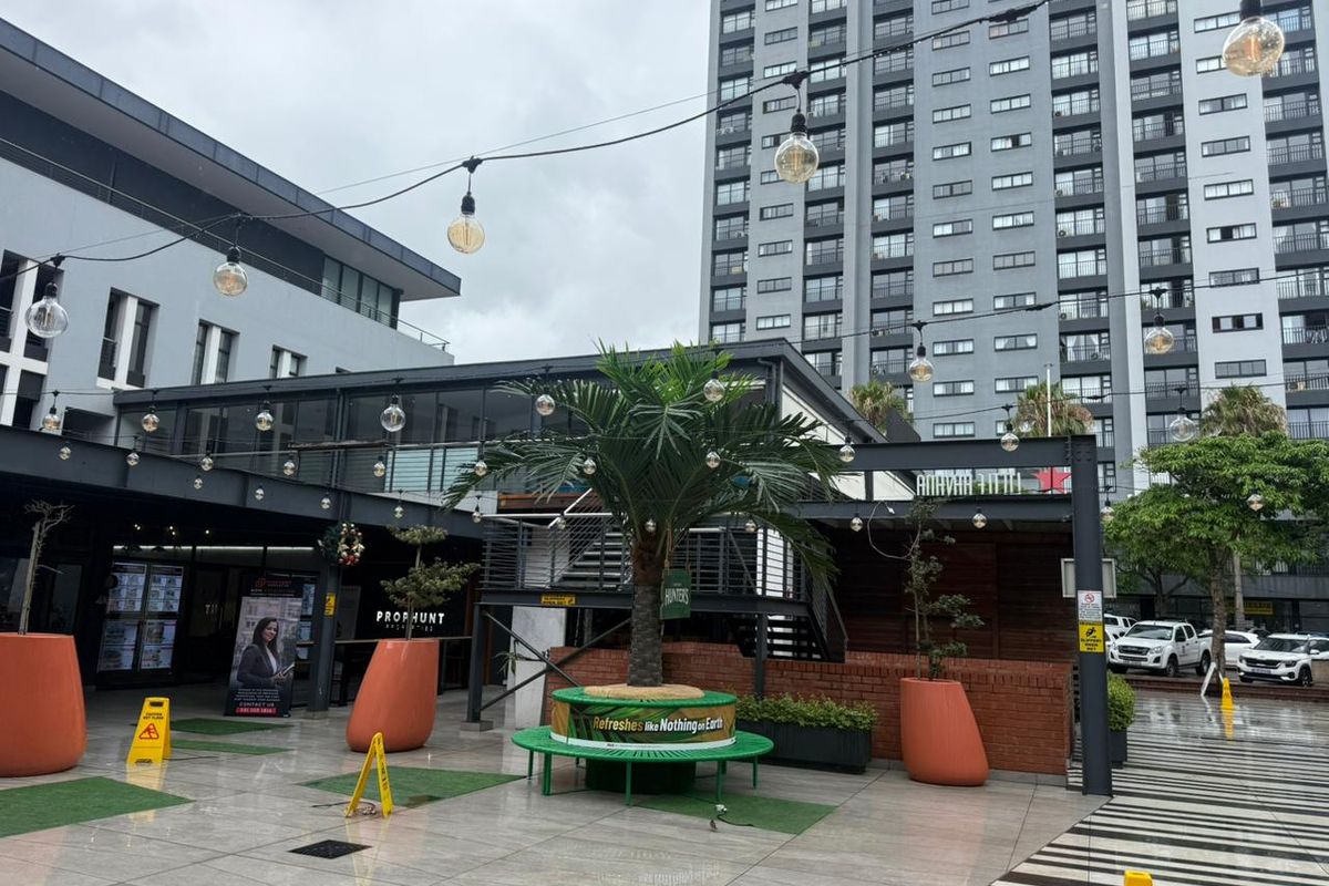 An outdoor retail courtyard area featuring paved flooring, overhead string lighting, and a central seating area with integrated landscaping.