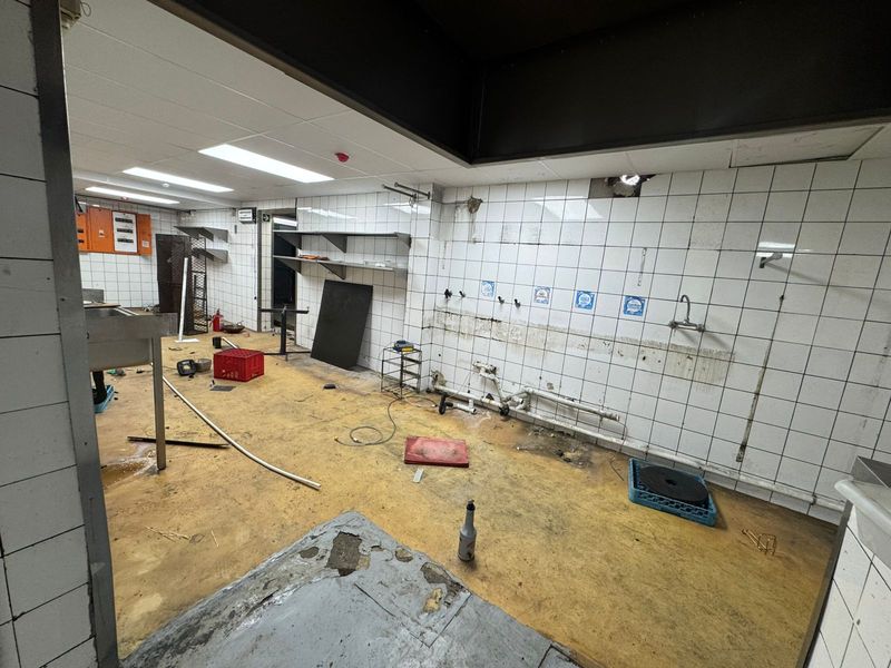 A vacant commercial kitchen space featuring tiled walls, exposed plumbing, and worn flooring, requiring significant renovation and refurbishment.