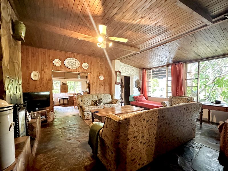 Rustic living room featuring a stone fireplace, timber-clad walls and ceiling, stone flooring, and large windows providing natural light.