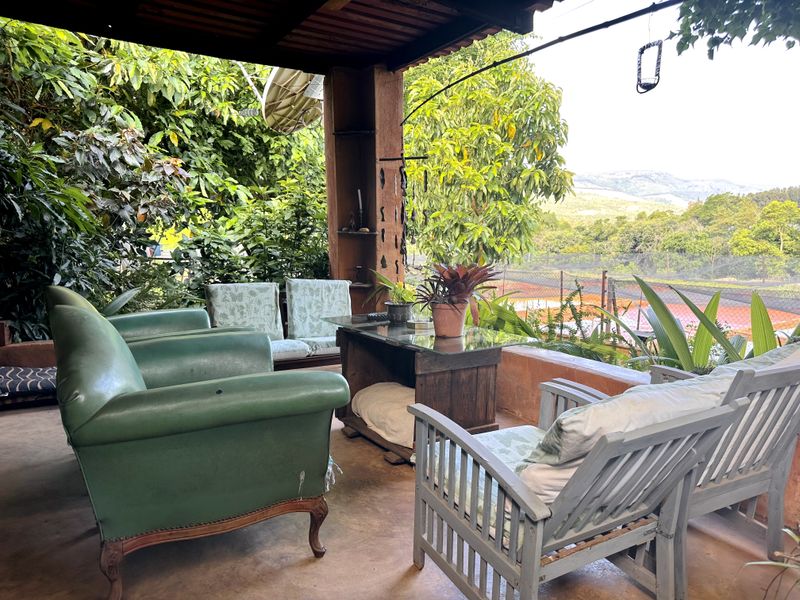 A covered outdoor patio area featuring concrete flooring and timber structural supports, overlooking a landscaped garden and tennis court.