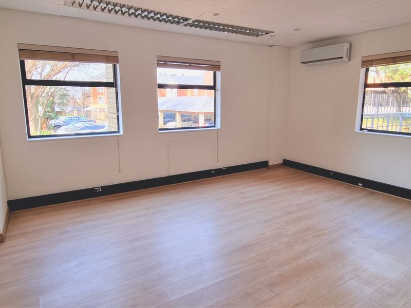 A bright, empty office space featuring laminate flooring, recessed lighting, and a wall-mounted air conditioning unit with large windows providing natural light.