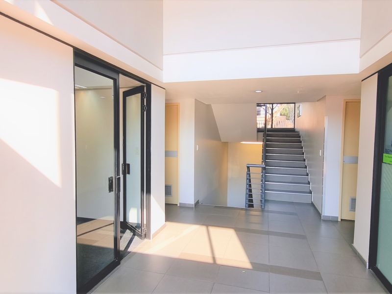 A bright and modern office building entrance featuring tiled flooring, glass partition doors, and a central staircase leading to upper levels.