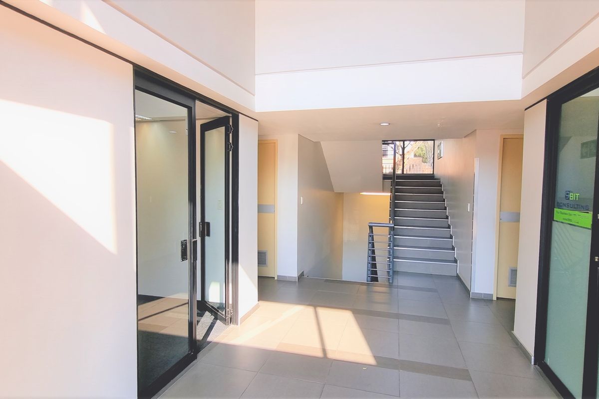 A bright and modern office building entrance featuring tiled flooring, glass partition doors, and a central staircase leading to upper levels.