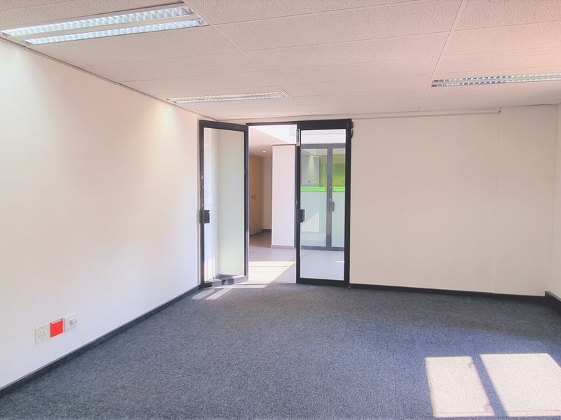 A bright, empty office space featuring carpeted flooring, a suspended ceiling with recessed lighting, and glass entry doors.