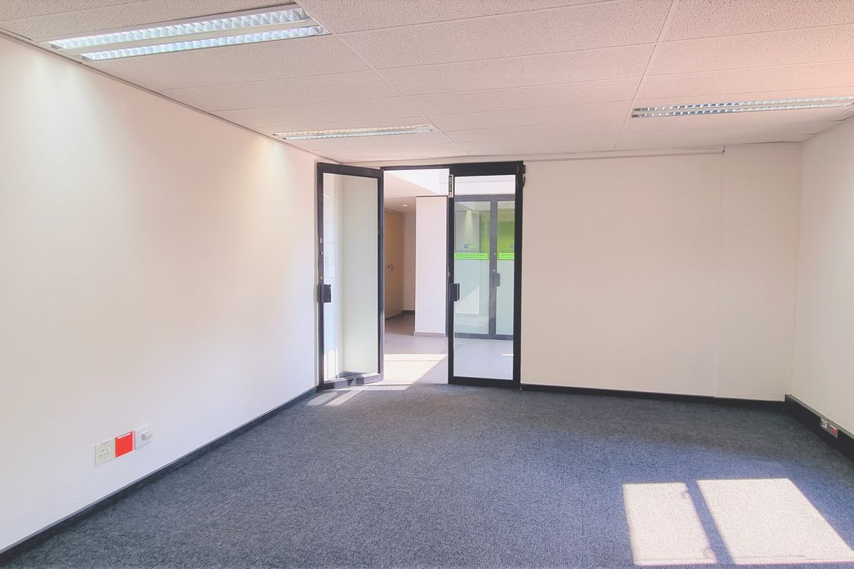 A bright, empty office space featuring carpeted flooring, a suspended ceiling with recessed lighting, and glass entry doors.