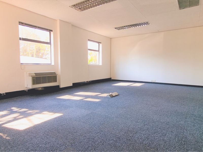 A spacious, empty office area featuring carpeted flooring, recessed ceiling lighting, and a wall-mounted air conditioning unit.