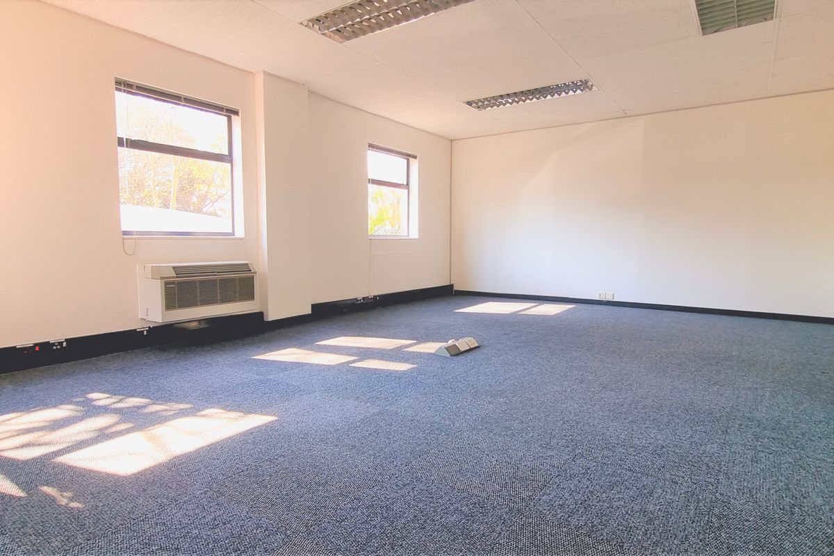 A spacious, empty office area featuring carpeted flooring, recessed ceiling lighting, and a wall-mounted air conditioning unit.
