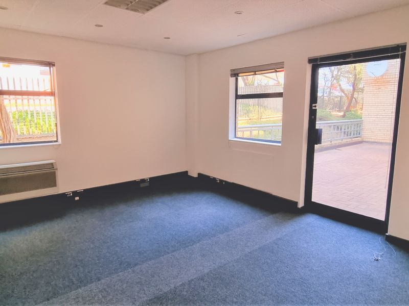 A bright, empty office space featuring carpeted flooring, a suspended ceiling with recessed lighting, an integrated HVAC unit, and direct access to an outdoor patio.