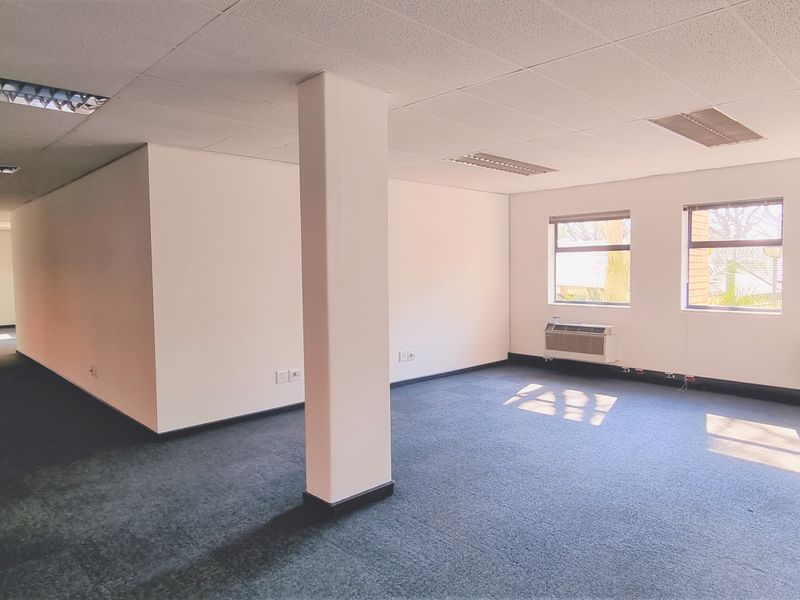 A spacious, empty open-plan office area featuring carpeted flooring, suspended ceilings with recessed lighting, and multiple windows providing natural light.