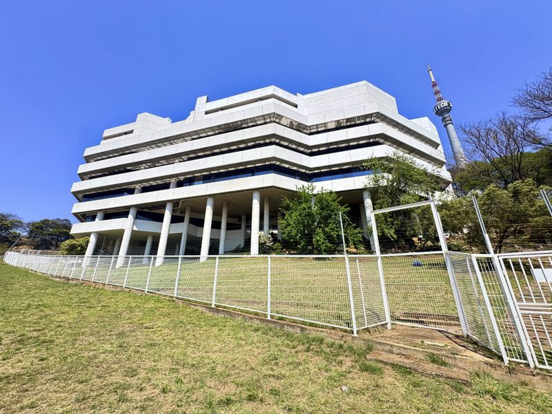 A multi-level office building featuring a distinctive concrete facade, glass windows, and structural pillars, secured by perimeter fencing.