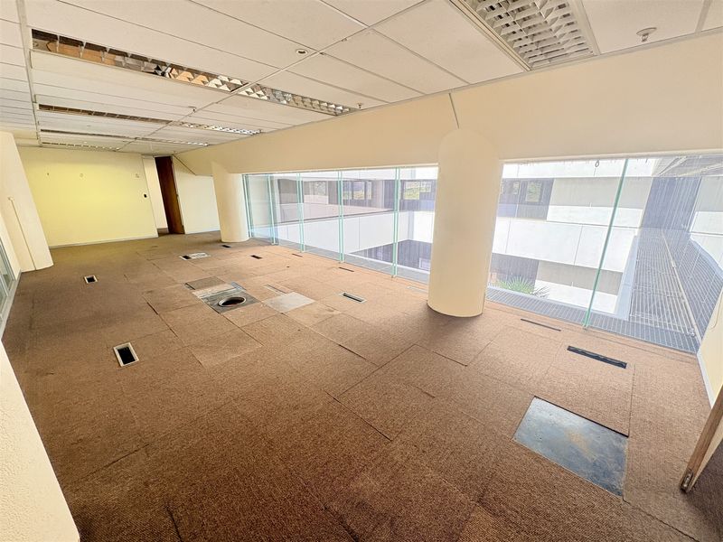 An empty, open-plan office space featuring raised access flooring, suspended ceilings with recessed lighting, and large glass windows overlooking an internal courtyard.