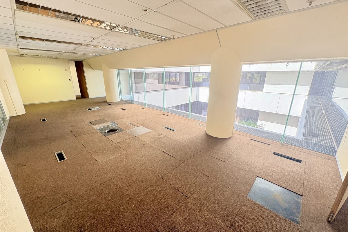 An empty, open-plan office space featuring raised access flooring, suspended ceilings with recessed lighting, and large glass windows overlooking an internal courtyard.