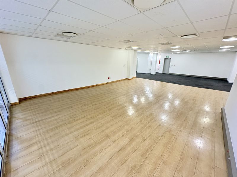 An open-plan office space featuring a mix of laminate and carpet flooring, suspended ceilings with recessed lighting, and a neutral white finish.