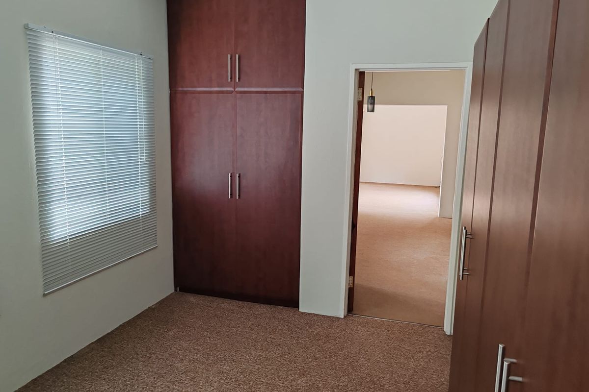 A bedroom featuring built-in wooden cabinetry, carpeted flooring, and a window with fitted blinds, leading into an adjacent room.