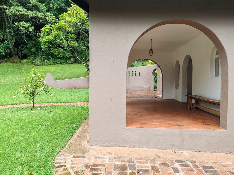 A covered patio area featuring classic arched architecture, terracotta tiled flooring, and direct access to a lush, well-maintained garden.