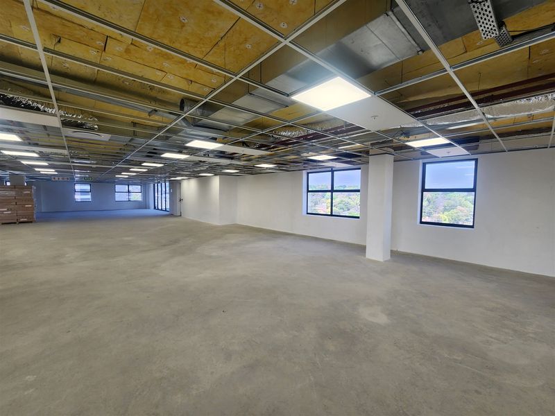 Spacious open-plan office area featuring an unfinished ceiling, exposed ducting, concrete flooring, and large windows providing ample natural light.