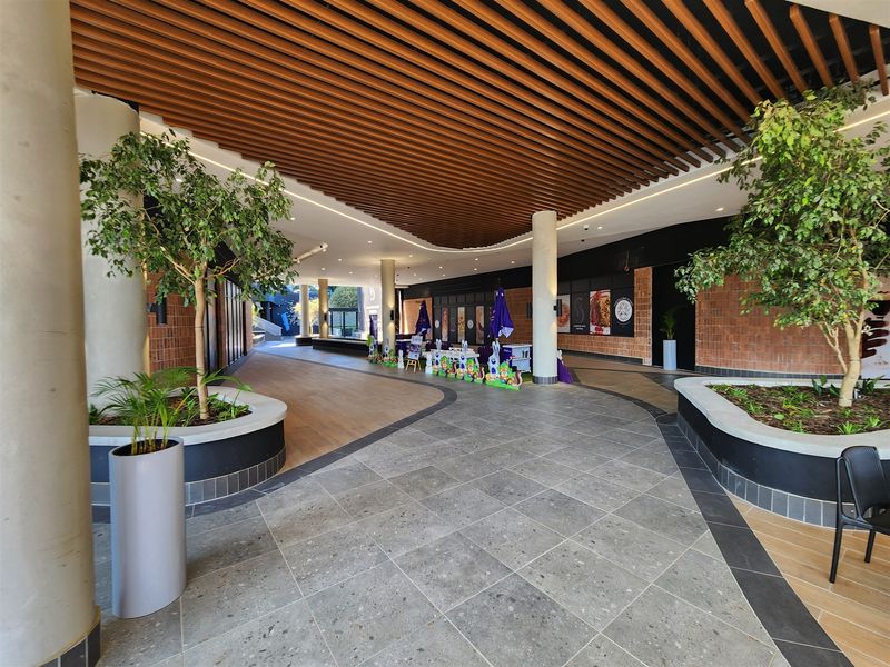 Modern office building entrance lobby featuring a slatted timber ceiling, concrete columns, and integrated planters with a mix of stone and wood-look flooring.