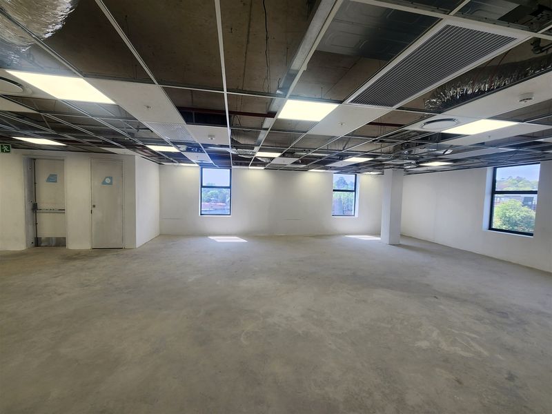 An open-plan office space featuring an exposed concrete ceiling, suspended grid lighting, and large windows, currently in a shell condition ready for fit-out.