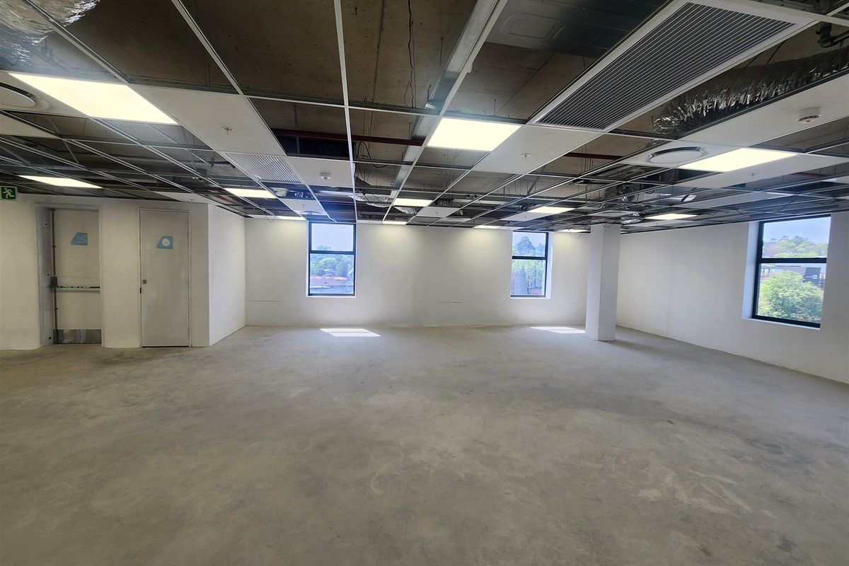 An open-plan office space featuring an exposed concrete ceiling, suspended grid lighting, and large windows, currently in a shell condition ready for fit-out.