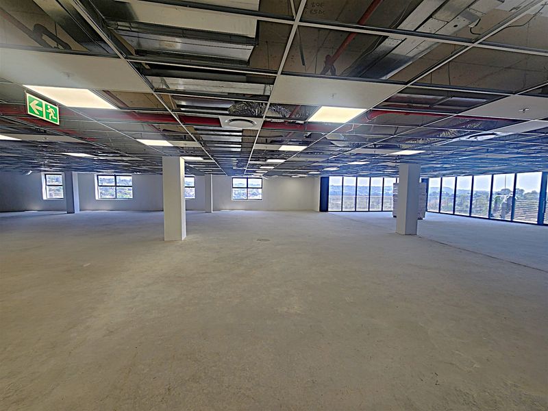 A large, open-plan office space currently under renovation, featuring an exposed ceiling grid, structural columns, and expansive windows with natural light.
