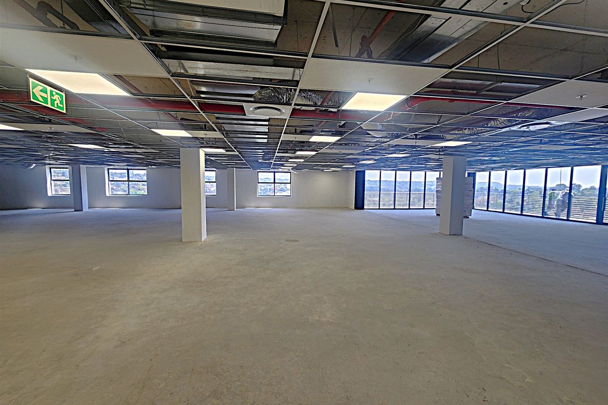 A large, open-plan office space currently under renovation, featuring an exposed ceiling grid, structural columns, and expansive windows with natural light.