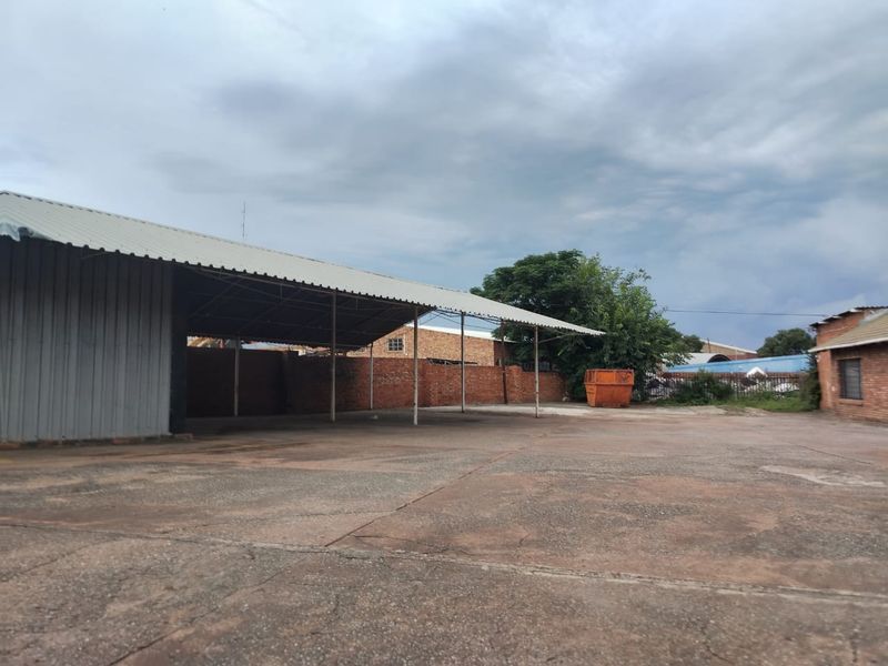 A large, open-air commercial yard featuring a covered steel-framed structure, brick boundary walls, and extensive concrete paving suitable for storage or parking.
