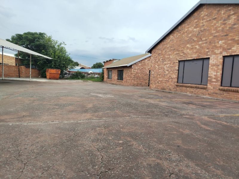 Exterior view of a commercial property featuring a large concrete paved yard, brick building facade, and a covered parking area.