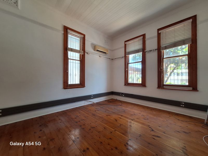A bright, empty office space featuring polished timber flooring, high ceilings, multiple windows with timber frames, and a wall-mounted air conditioning unit.