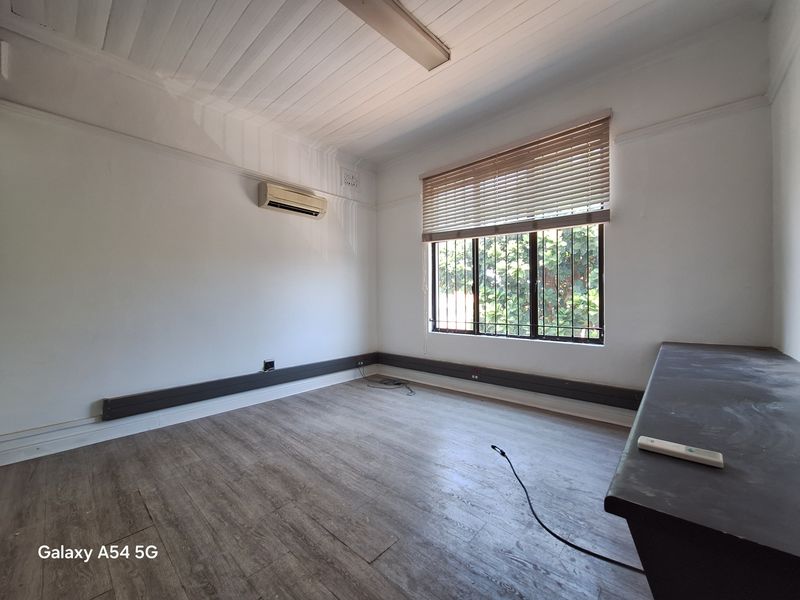 A functional office space featuring wood-look flooring, a wall-mounted air conditioning unit, and a window with fitted blinds and security bars.