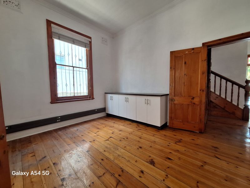 A bright office space featuring polished timber flooring, a built-in storage cabinet, and a window with security bars.