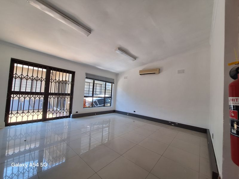 A bright, open-plan office space featuring polished tile flooring, security gates, and a wall-mounted air conditioning unit.