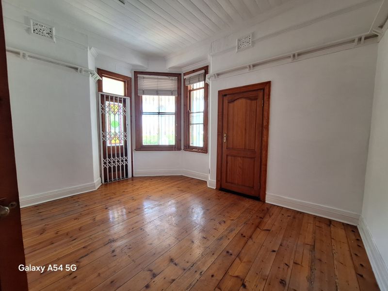A bright, empty office space featuring polished timber flooring, classic timber door, and a bay window with security bars.