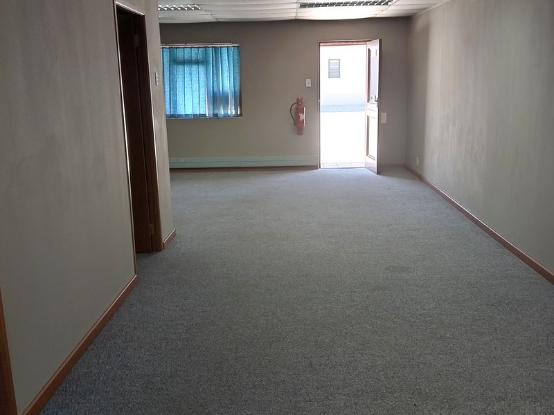An empty, open-plan office space featuring a suspended ceiling with integrated lighting, carpeted flooring, and a wall-mounted fire extinguisher.