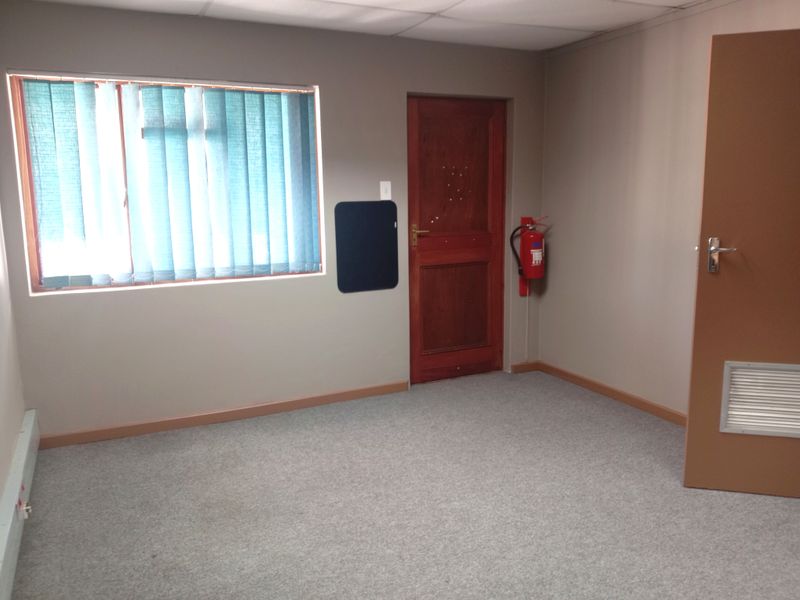 A functional office space featuring neutral walls, carpeted flooring, a wooden door, and a wall-mounted fire extinguisher.