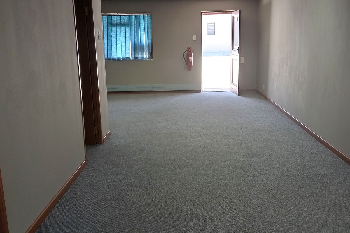 An empty, open-plan office space featuring a suspended ceiling with integrated lighting, carpeted flooring, and a wall-mounted fire extinguisher.