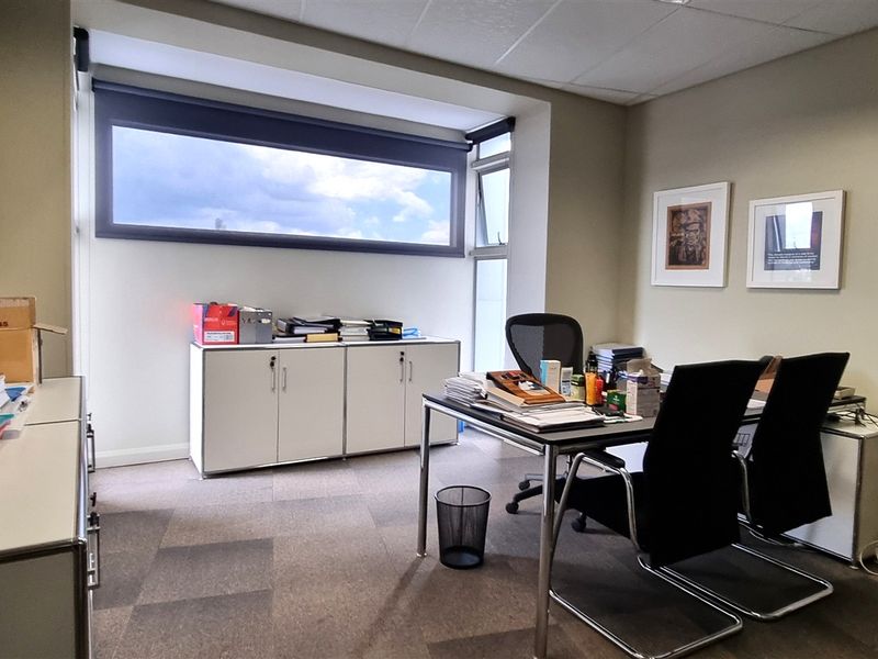 A functional office space featuring carpeted flooring, a suspended ceiling, and a horizontal window with a roller blind, providing natural light.