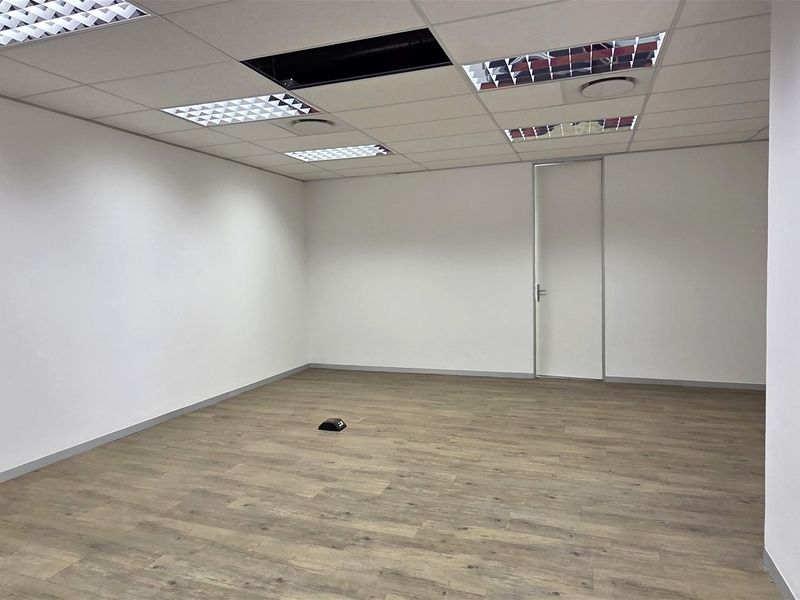 A vacant office space featuring laminate flooring, a suspended ceiling with recessed lighting, and a missing ceiling tile.