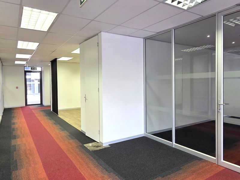 A bright office hallway featuring glass partition walls, suspended ceilings with recessed lighting, and patterned carpet tiles throughout the corridor.