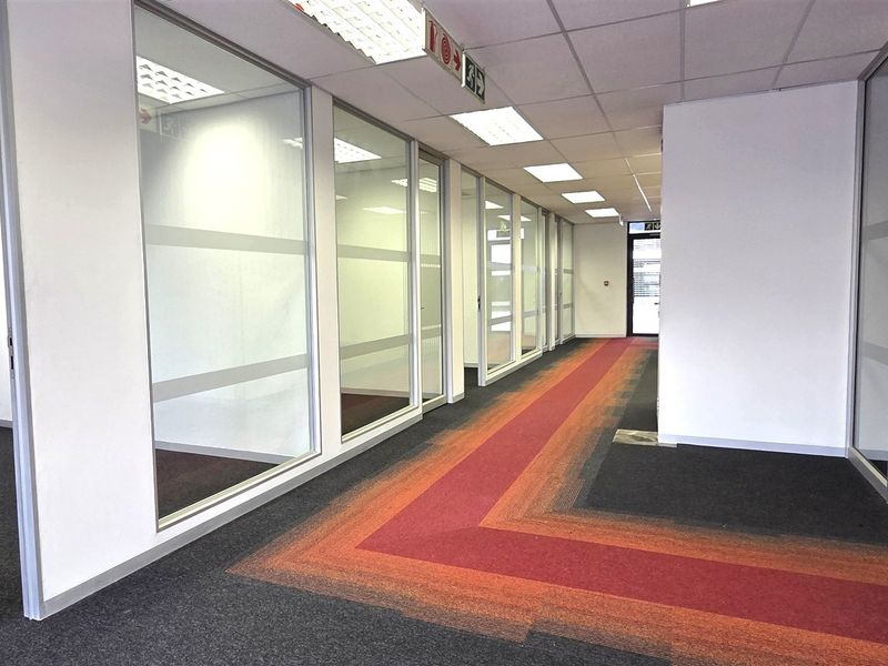 A modern office hallway featuring glass partition walls, suspended ceilings with recessed lighting, and patterned carpet tiles, leading to multiple private office spaces.