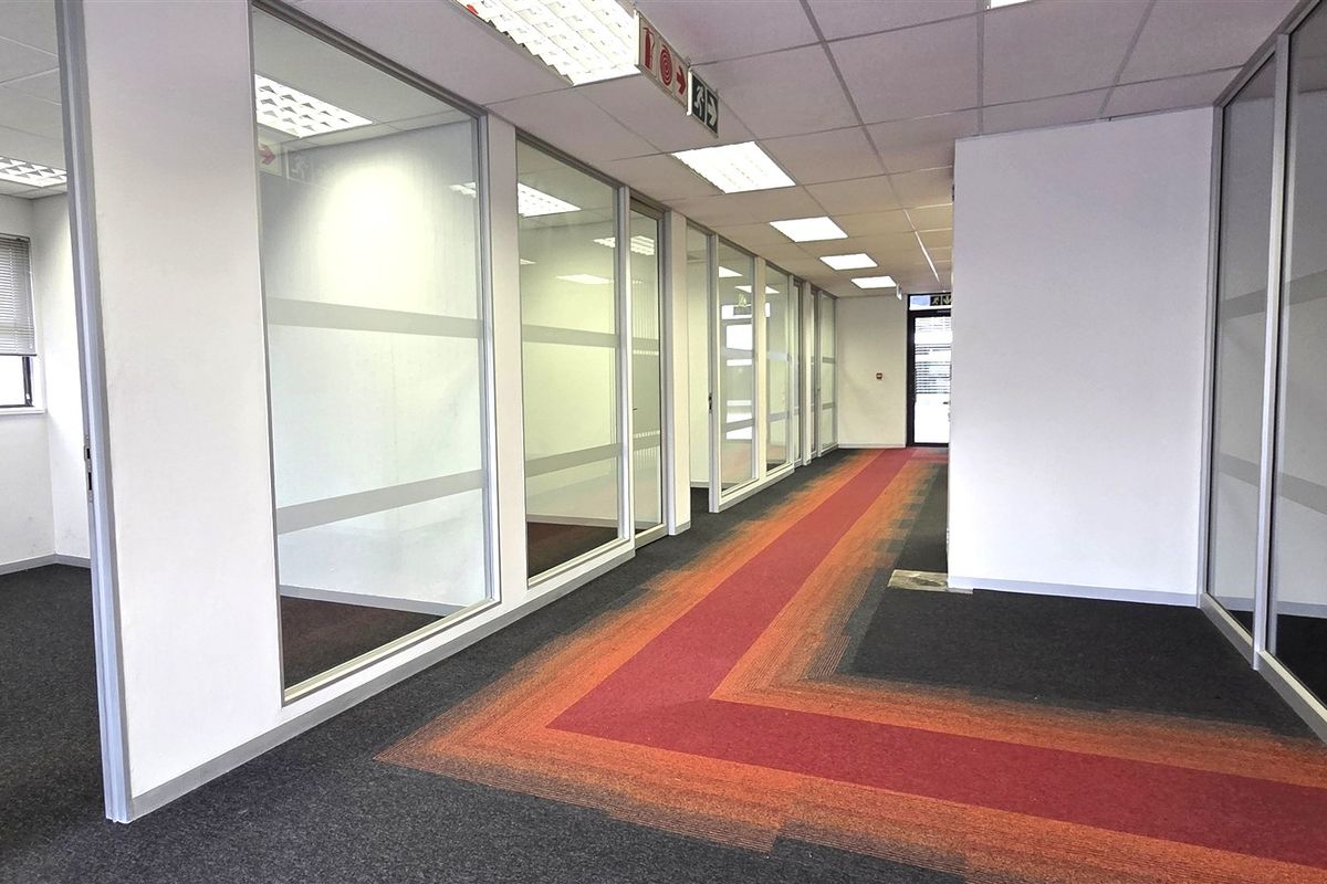 A modern office hallway featuring glass partition walls, suspended ceilings with recessed lighting, and patterned carpet tiles, leading to multiple private office spaces.