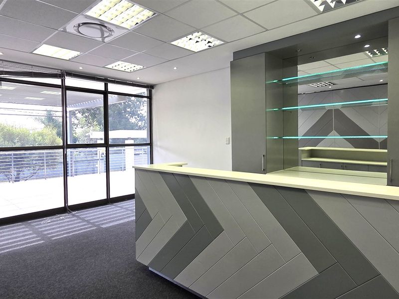 Modern office space featuring a custom built-in reception desk, sleek cabinetry, suspended ceiling with recessed lighting, and large glass doors leading to an outdoor area.