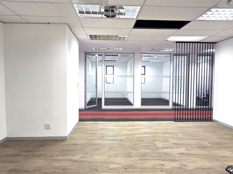 An empty office space featuring a mix of laminate and carpet flooring, glass partitioning, and a suspended ceiling with a missing tile.