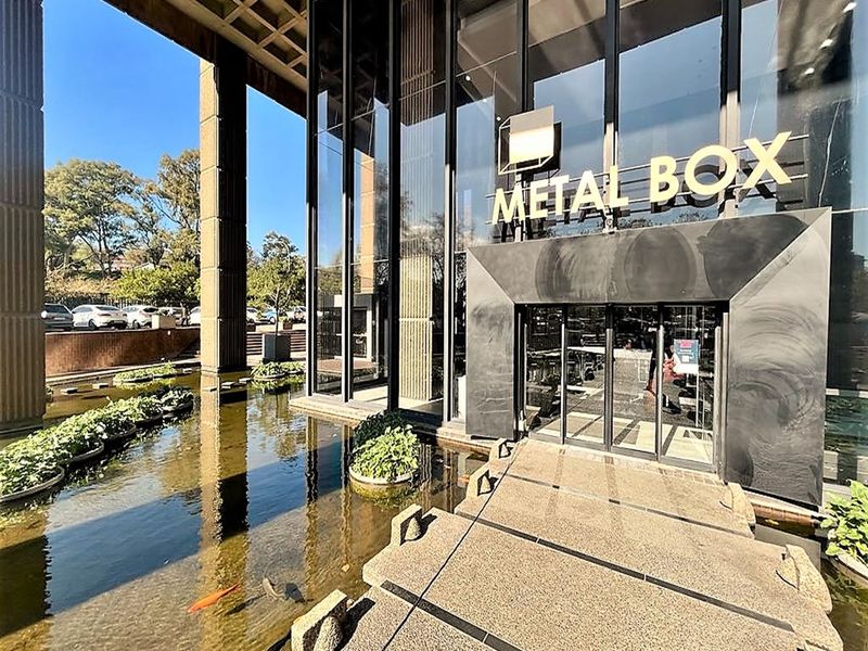 Modern office building entrance featuring a glass facade, concrete structural columns, and a decorative water feature with a stone pedestrian walkway.
