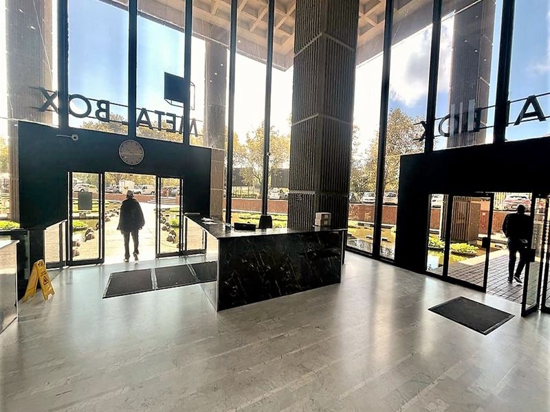 Spacious office building entrance lobby featuring high ceilings, floor-to-ceiling windows, marble reception desk, and modern stone flooring.