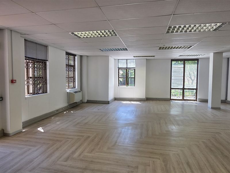 Spacious, open-plan office area featuring modern herringbone vinyl flooring, suspended ceilings with recessed lighting, and large windows with security gates.