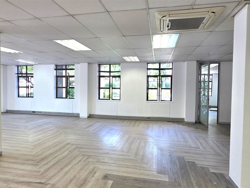 Spacious open-plan office area featuring modern herringbone flooring, suspended ceilings with integrated lighting, cassette air conditioning, and large windows providing ample natural light.