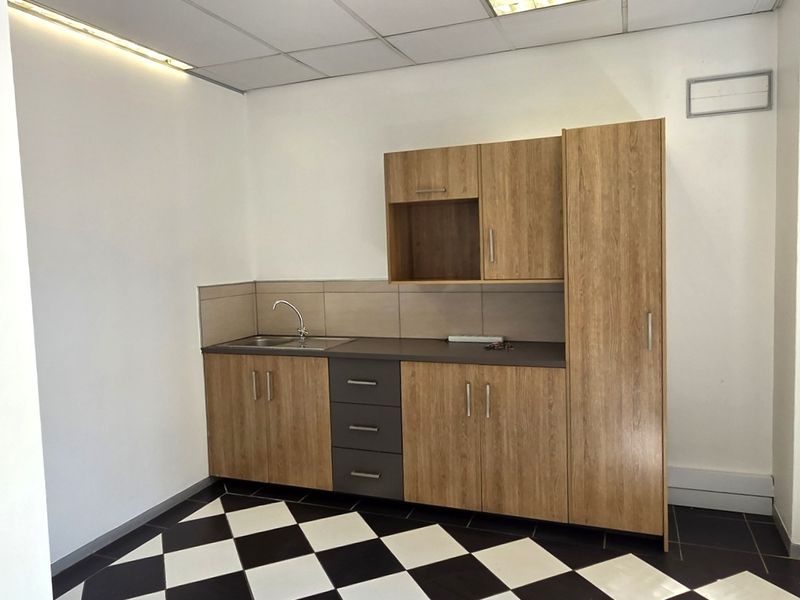 A clean, compact office kitchenette featuring wood-look cabinetry, a stainless steel sink, tiled backsplash, and a modern black and white checkered floor.
