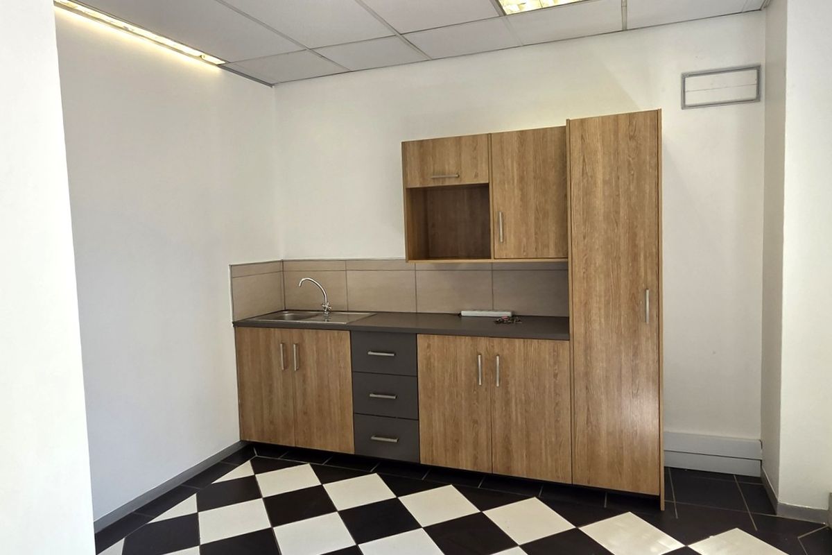 A clean, compact office kitchenette featuring wood-look cabinetry, a stainless steel sink, tiled backsplash, and a modern black and white checkered floor.